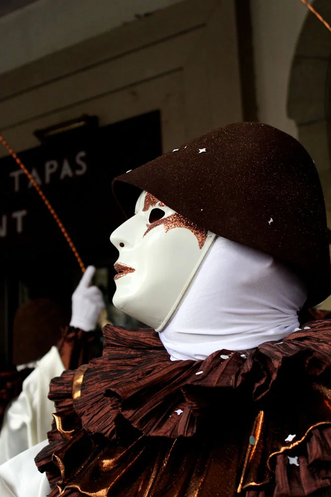 Side-profile portrait of a masked Les Rambailurs performer in ivory and white with a copper-brown ruff and dark Pierrot hat.